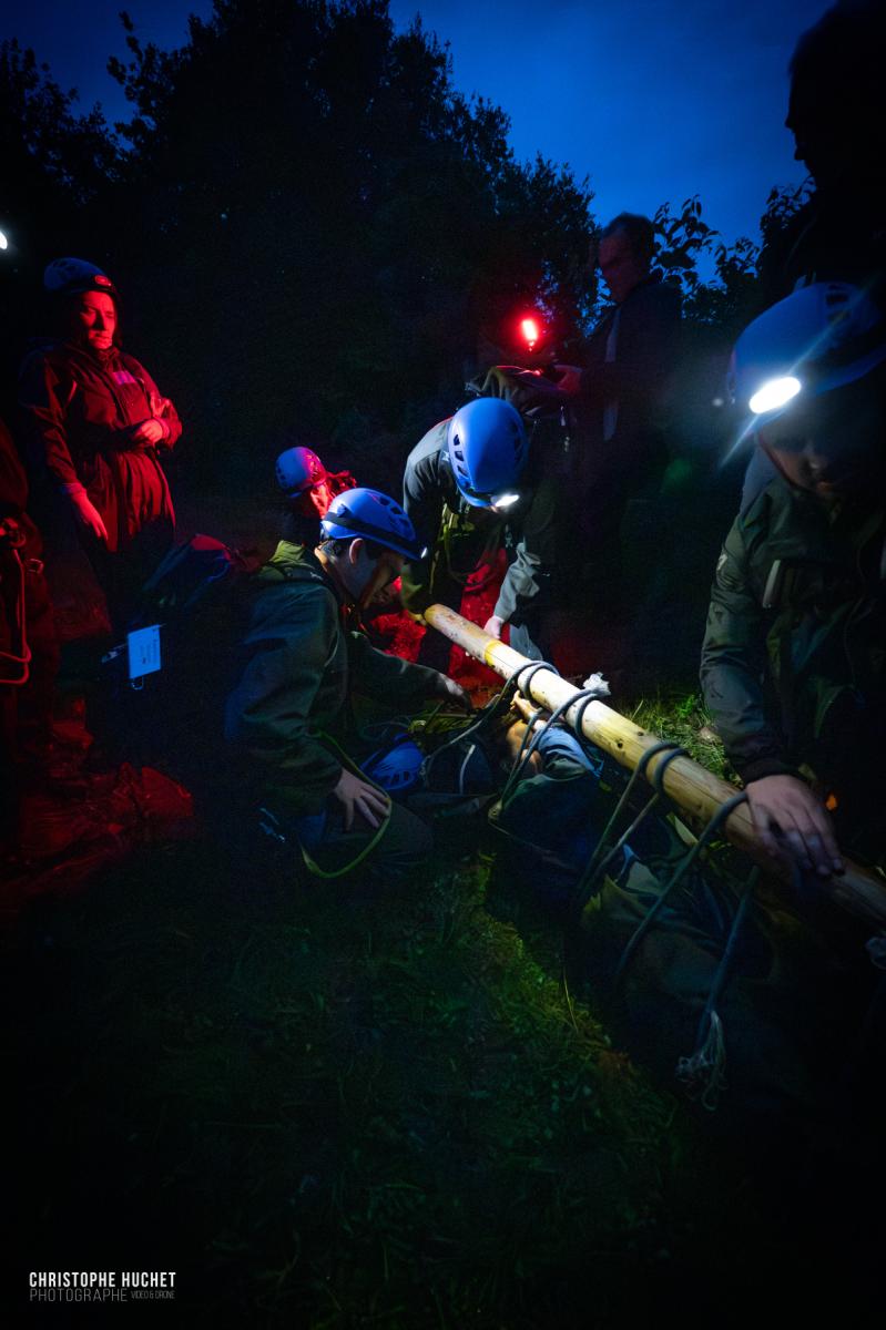 Équipe casquée avec lampes frontales manœuvrant un brancard de fortune lors d’un exercice nocturne en forêt.