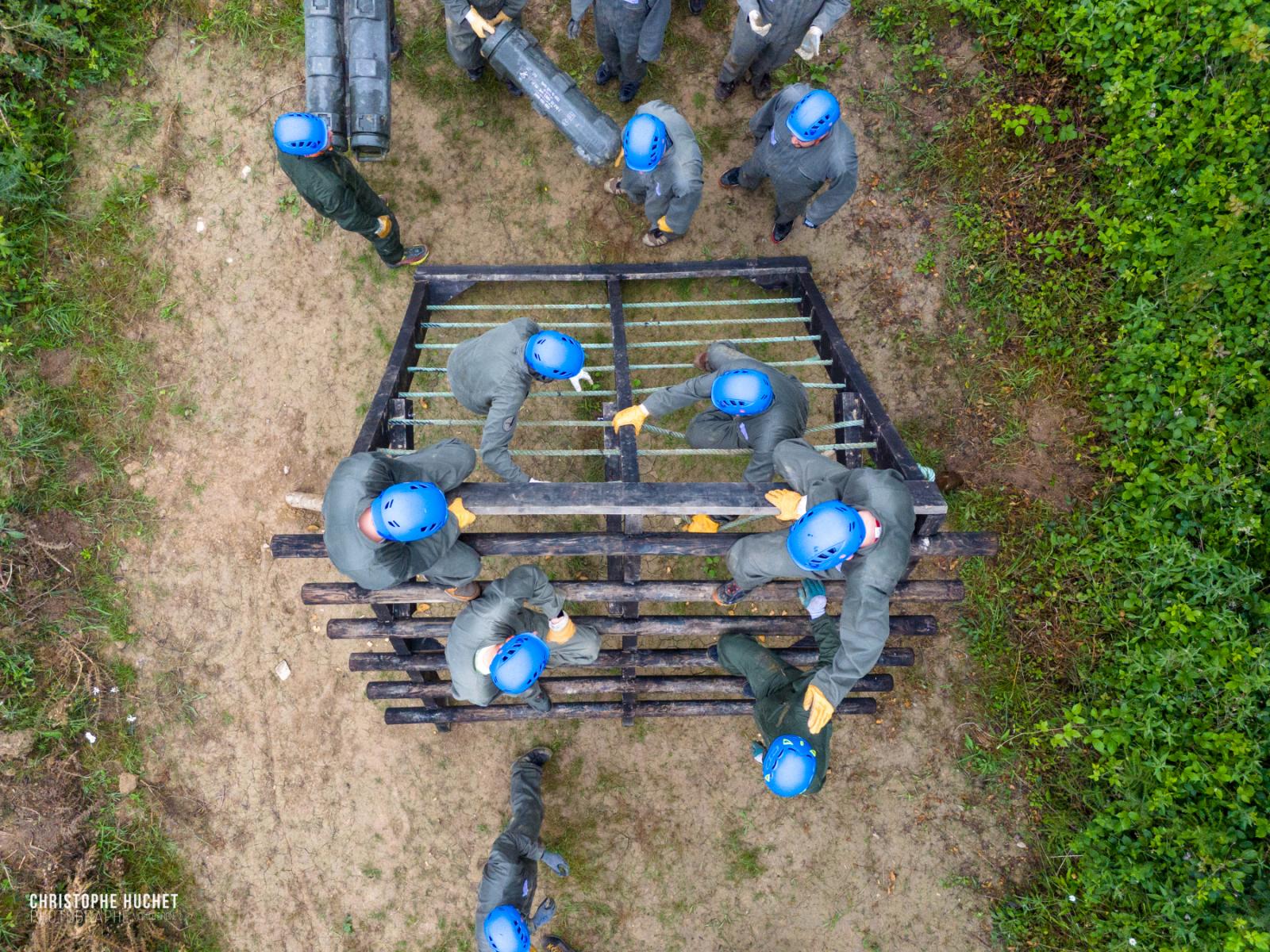 Vue drone de participants casqués franchissant un obstacle en bois lors d’un stage de cohésion d’équipe.