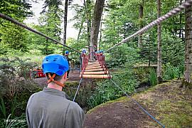 Homme au casque bleu observe des participants traversant un pont de cordes et bois dans une forêt lumineuse en Bretagne.