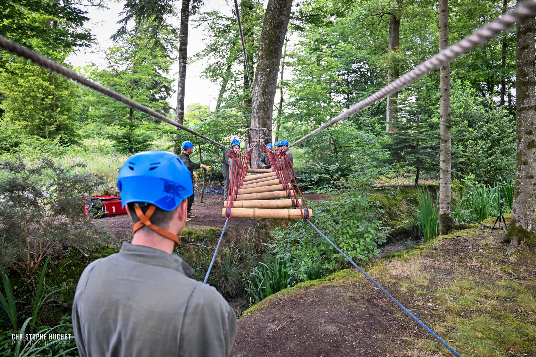 Homme au casque bleu observe des participants traversant un pont de cordes et bois dans une forêt lumineuse en Bretagne.