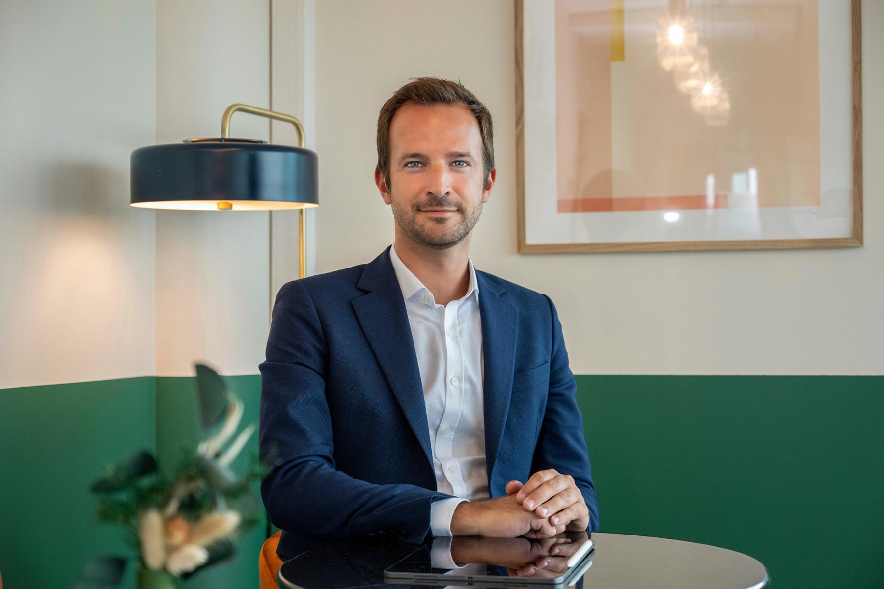 Homme jeune en costume bleu assis à un bureau moderne avec lampe design et cadre artistique, lumière naturelle douce