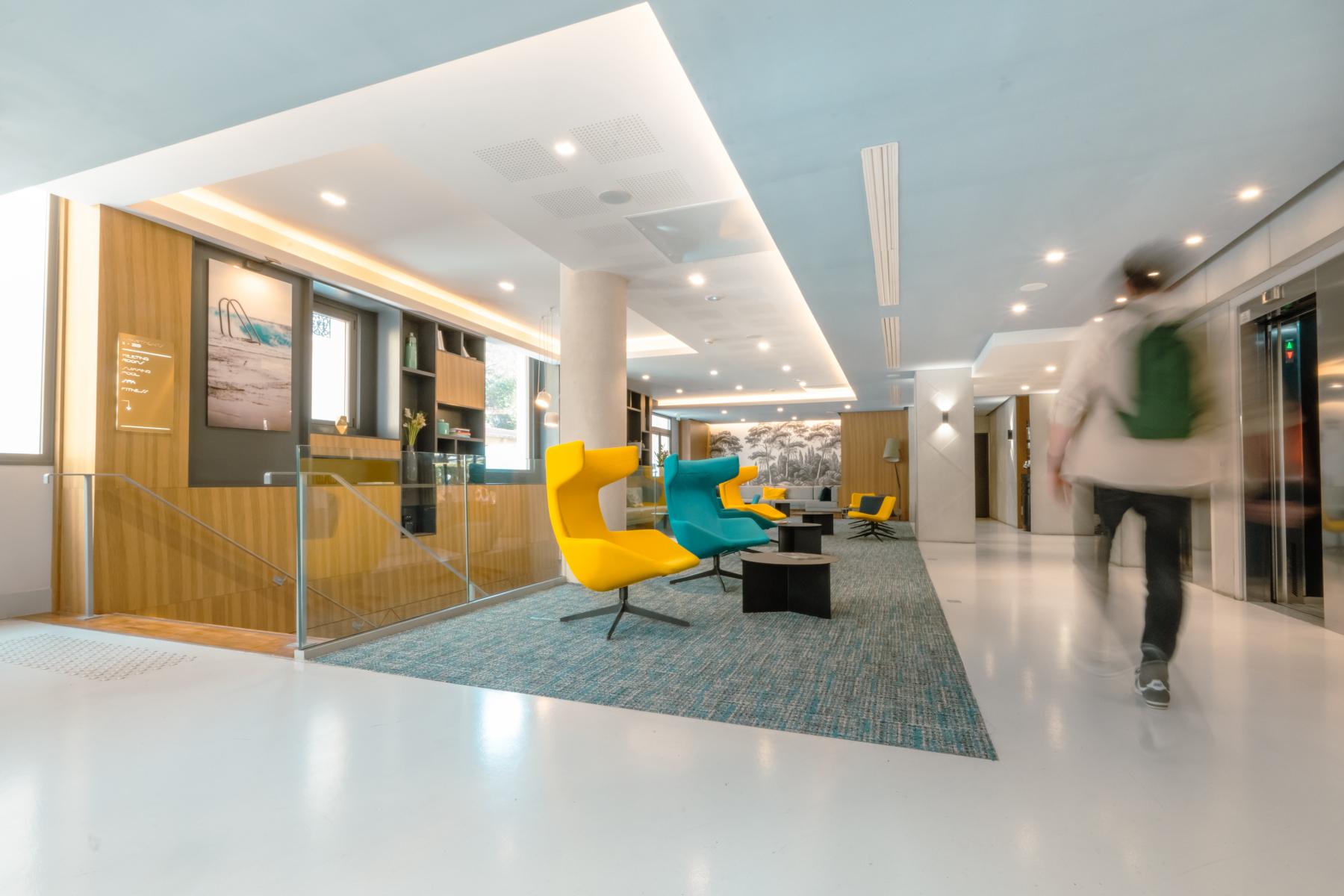 Hall d’hôtel à Cannes avec fauteuils jaune et bleu, sol blanc brillant et éclairage moderne au plafond.