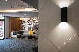 Applique murale noire sur mur texturé ouvrant sur un salon d’hôtel design avec fresque murale de forêt.