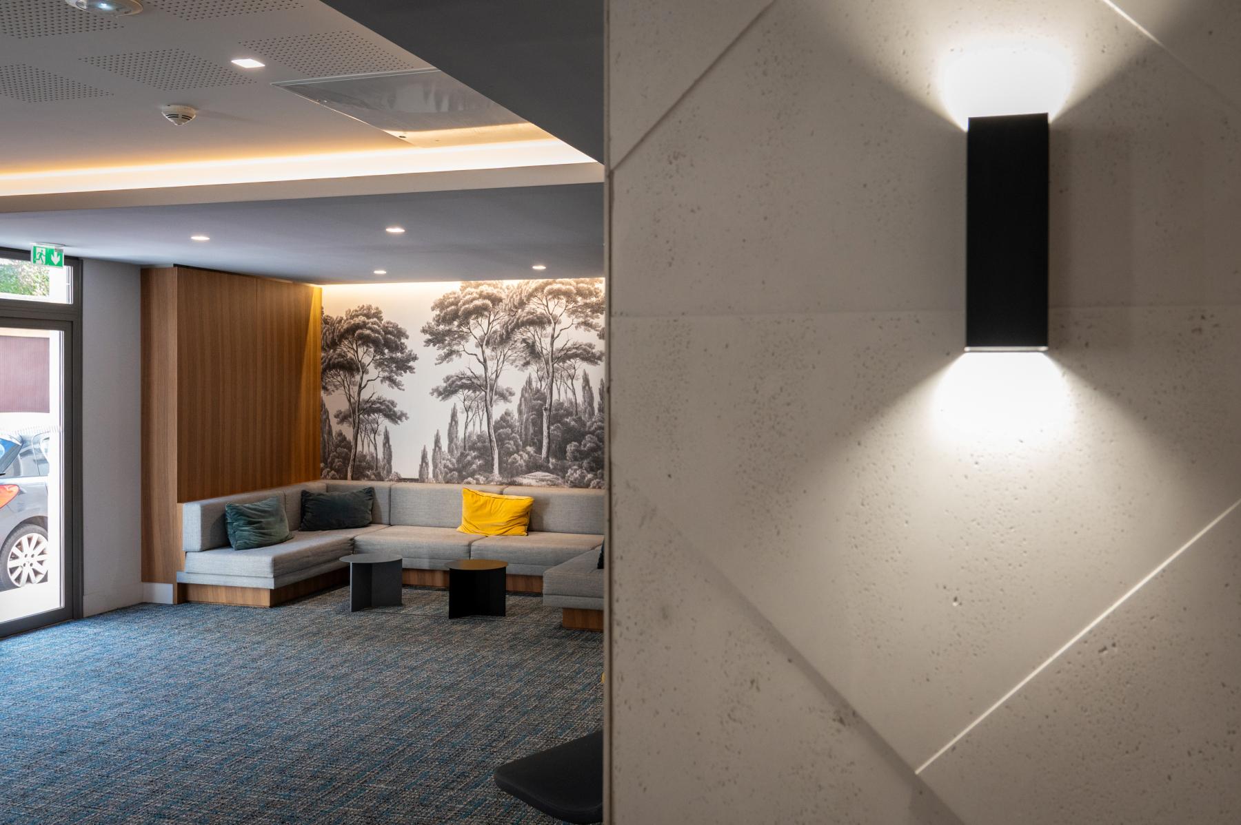Applique murale noire sur mur texturé ouvrant sur un salon d’hôtel design avec fresque murale de forêt.