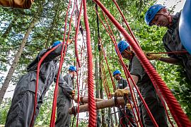 Équipe en casque bleu et combinaison kaki manipulant des cordes rouges et des rondins de bois pour la construction d’un pont dans une fôret.