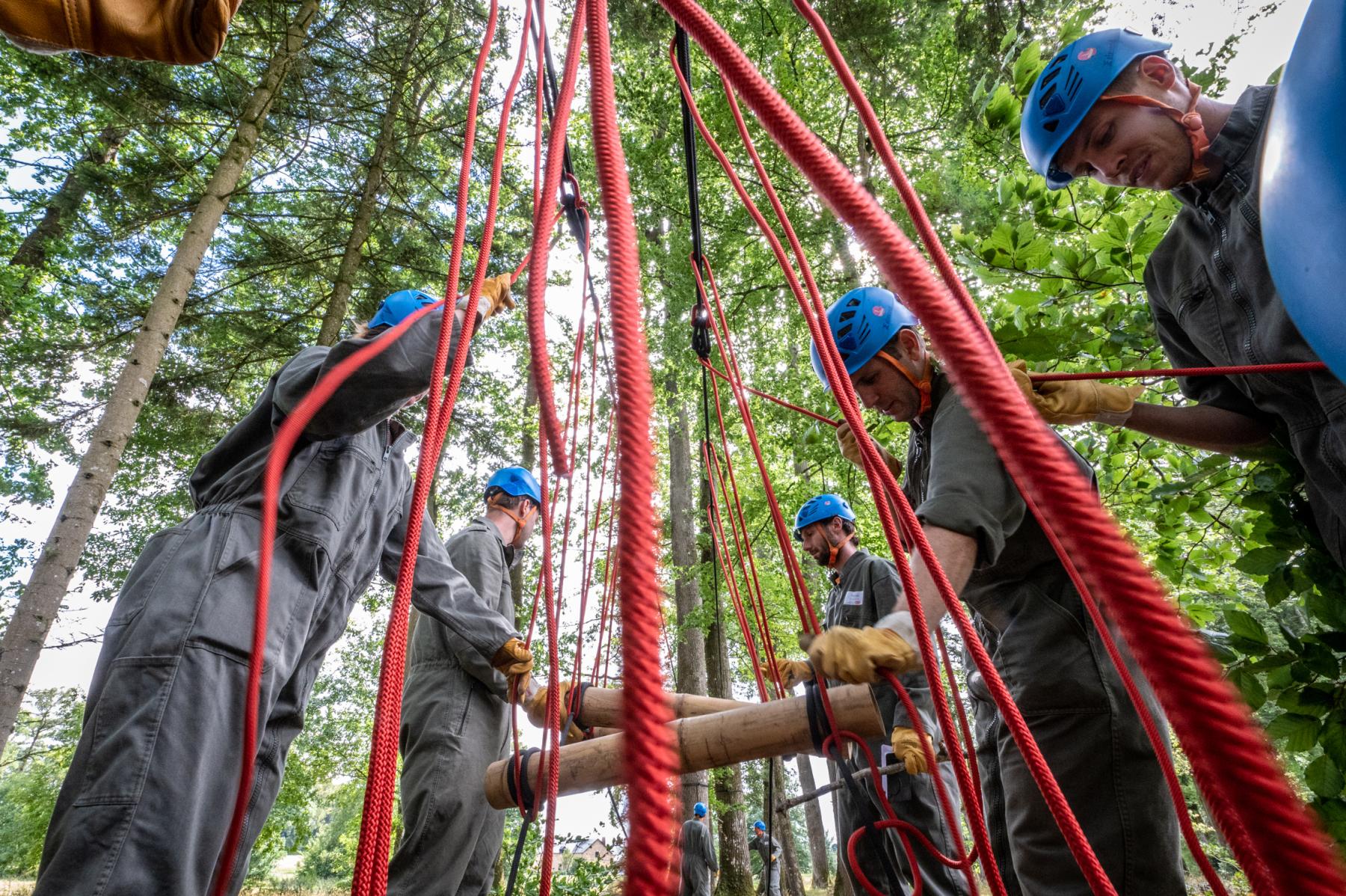 Équipe en casque bleu et combinaison kaki manipulant des cordes rouges et des rondins de bois pour la construction d’un pont dans une fôret.