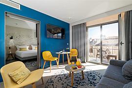 Salon d’appart-hôtel avec mur bleu, fauteuils jaunes et balcon ensoleillé offrant une vue sur la chambre.