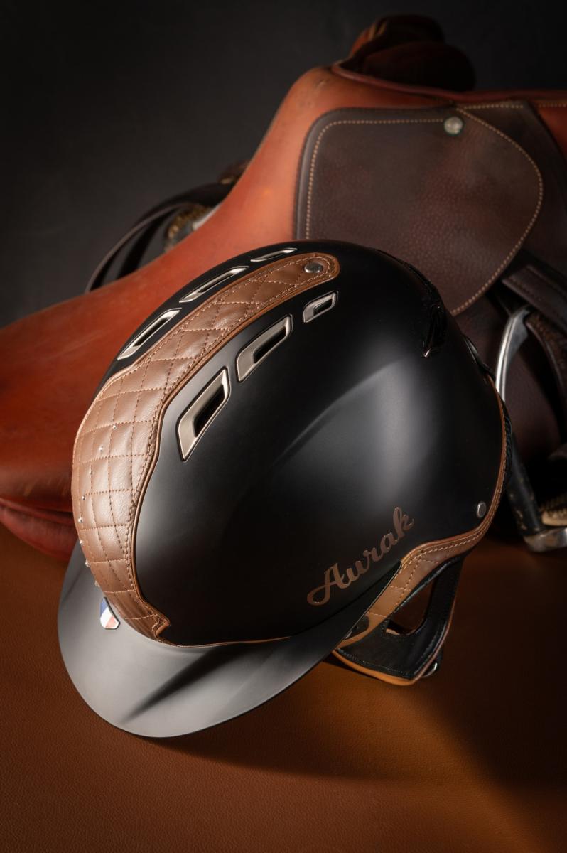 Casque d’équitation noir mat et cuir surpiqué devant une selle ancienne. Éclairage studio contrasté sur les textures cuir.