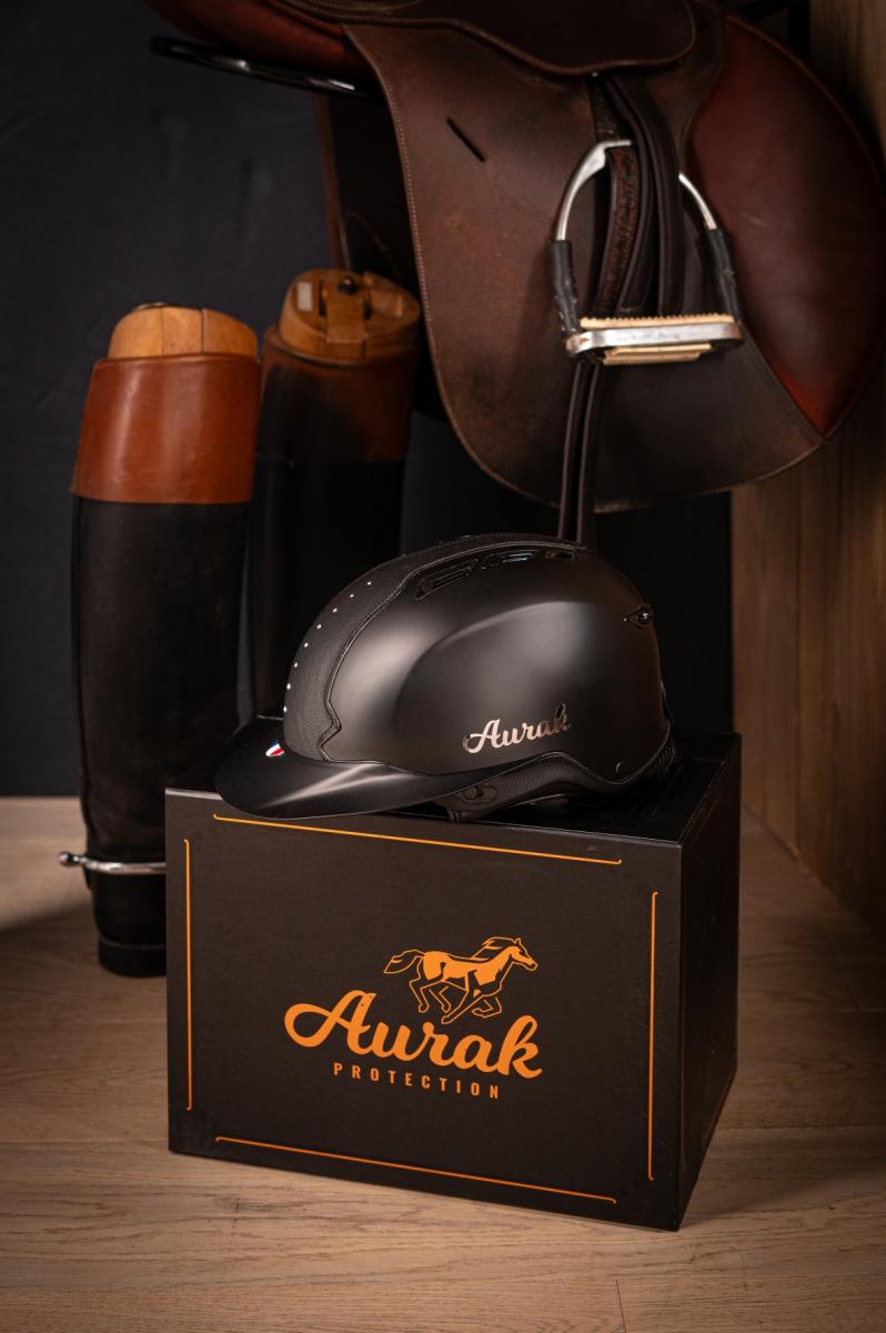 Casque équitation noir mat Aurak sur boîte, entouré de bottes et selle en cuir sous un éclairage studio précis.