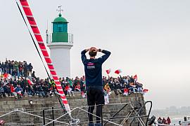 Skipper de dos sur son voilier saluant le public sur la jetée des Sables d’Olonne devant le phare vert.