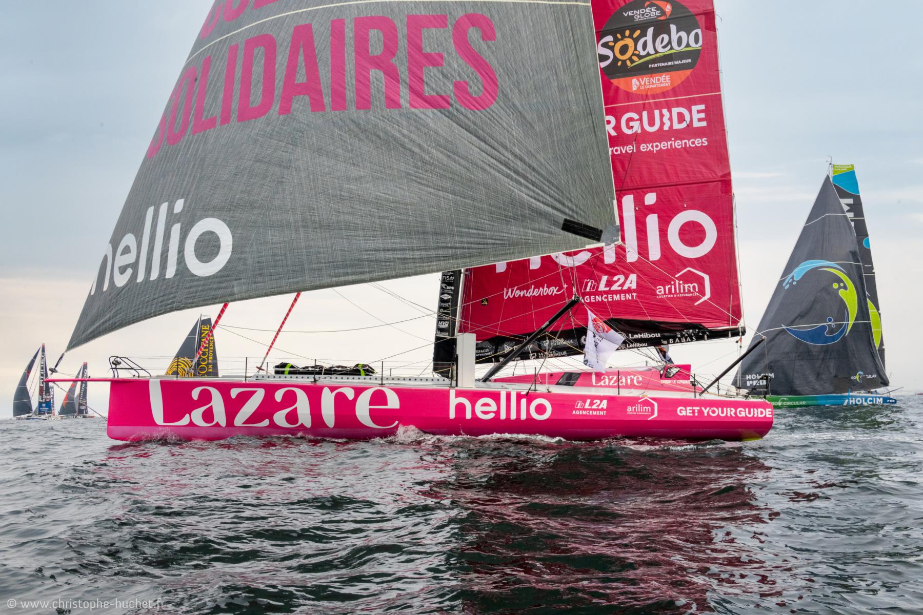 Voilier de course rose Lazare navigant avec ses voiles déployées lors du départ du Vendée Globe 2024 sur une mer calme.
