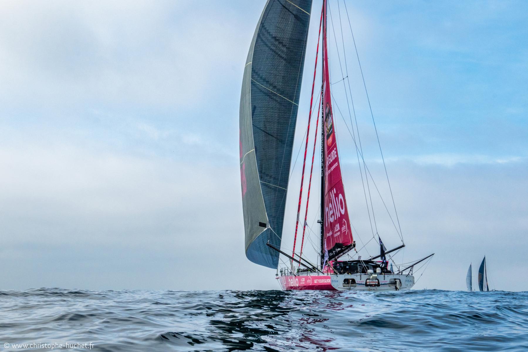 Voilier Imoca Lazare, voiles grises et roses sur mer formée, départ du Vendée Globe sous un ciel clair.