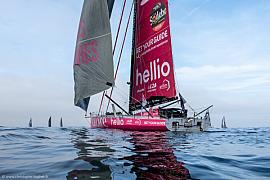 Voilier Imoca Lazare rose et blanc sur l’océan, grand-voile rouge Hellio sous un ciel voilé au départ de la régate.