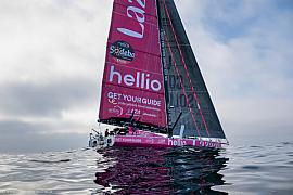 Voilier rose Imoca Lazare sur océan calme, ciel gris clair, reflets de l’eau, départ de course aux Sables-d’Olonne.