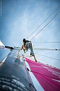 Marin grimpant au mât avec harnais, voile rose déployée sous un ciel bleu lors du départ du Vendée Globe sur l’Imoca Lazare.
