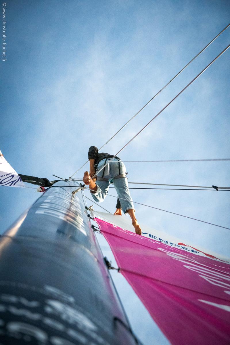 Marin grimpant au mât avec harnais, voile rose déployée sous un ciel bleu lors du départ du Vendée Globe sur l’Imoca Lazare.