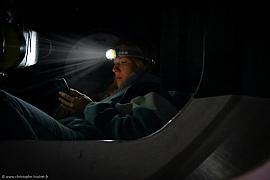 Portrait nocturne à la lampe frontale éclairant un smartphone dans la cabine de l’IMOCA Lazare.