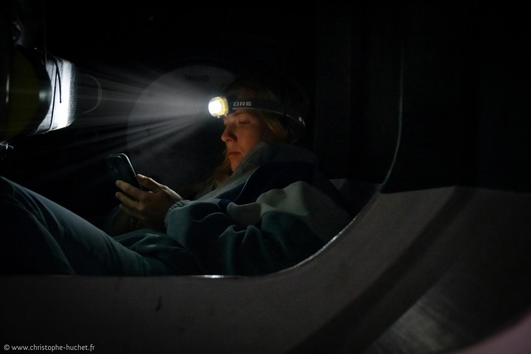 Portrait nocturne à la lampe frontale éclairant un smartphone dans la cabine de l’IMOCA Lazare.