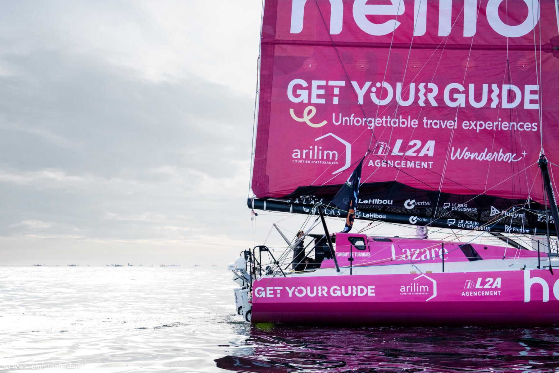Voilier IMOCA Lazare rose sur l’eau aux Sables d’Olonne, grand voile déployée sous un ciel nuageux.
