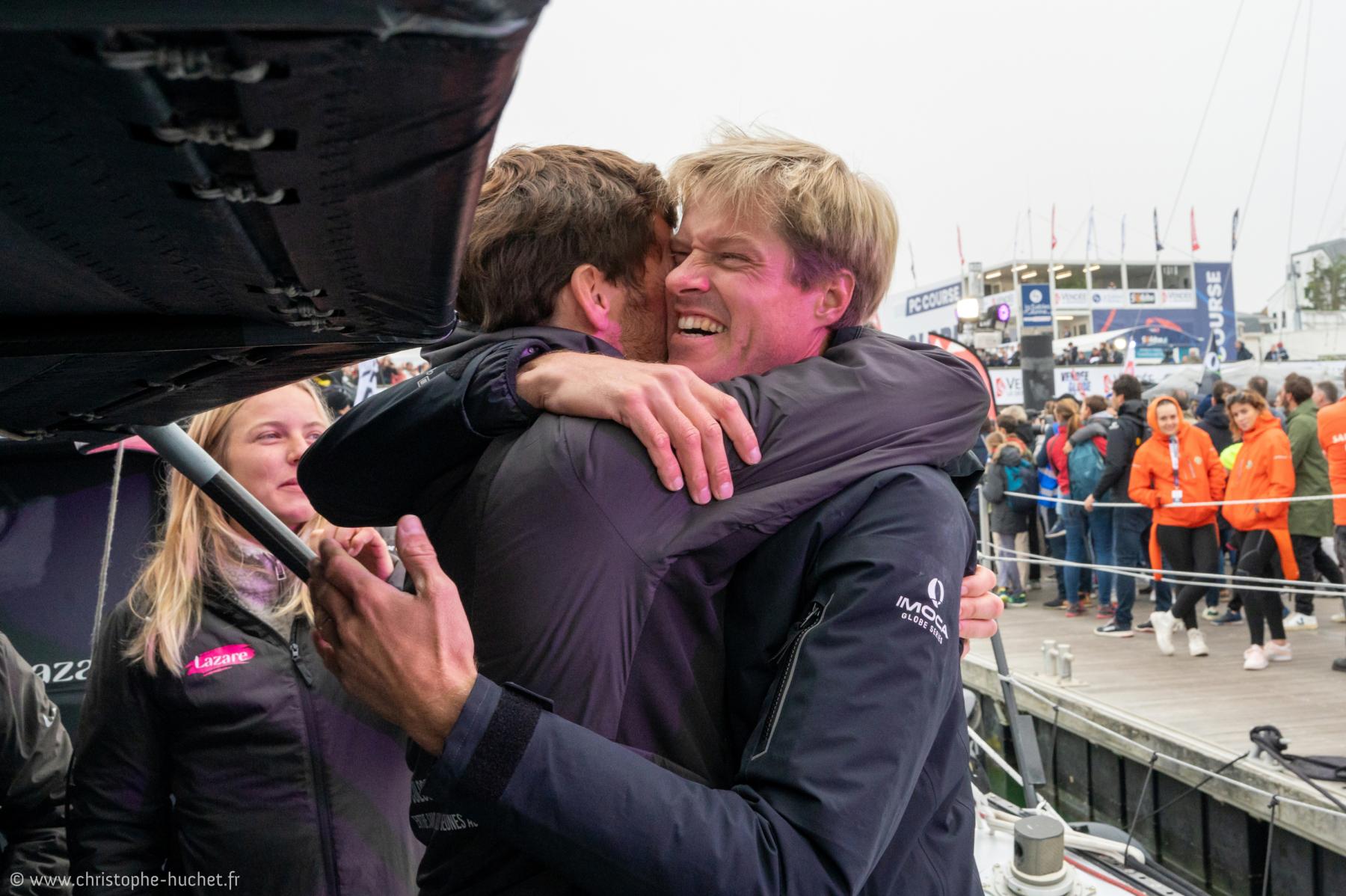 Étreinte émouvante entre deux skippers sur le pont d’un voilier IMOCA aux Sables-d’Olonne par temps gris.