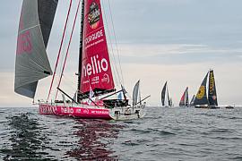 Voilier IMOCA Lazare rouge filant sur l’eau au départ du Vendée Globe 2024 aux Sables d’Olonne sous un ciel voilé.