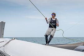 Marin sur un trimaran Ocean Fifty manipulant un cordage en mer sous un ciel clair lors d’une course à Saint-Malo.