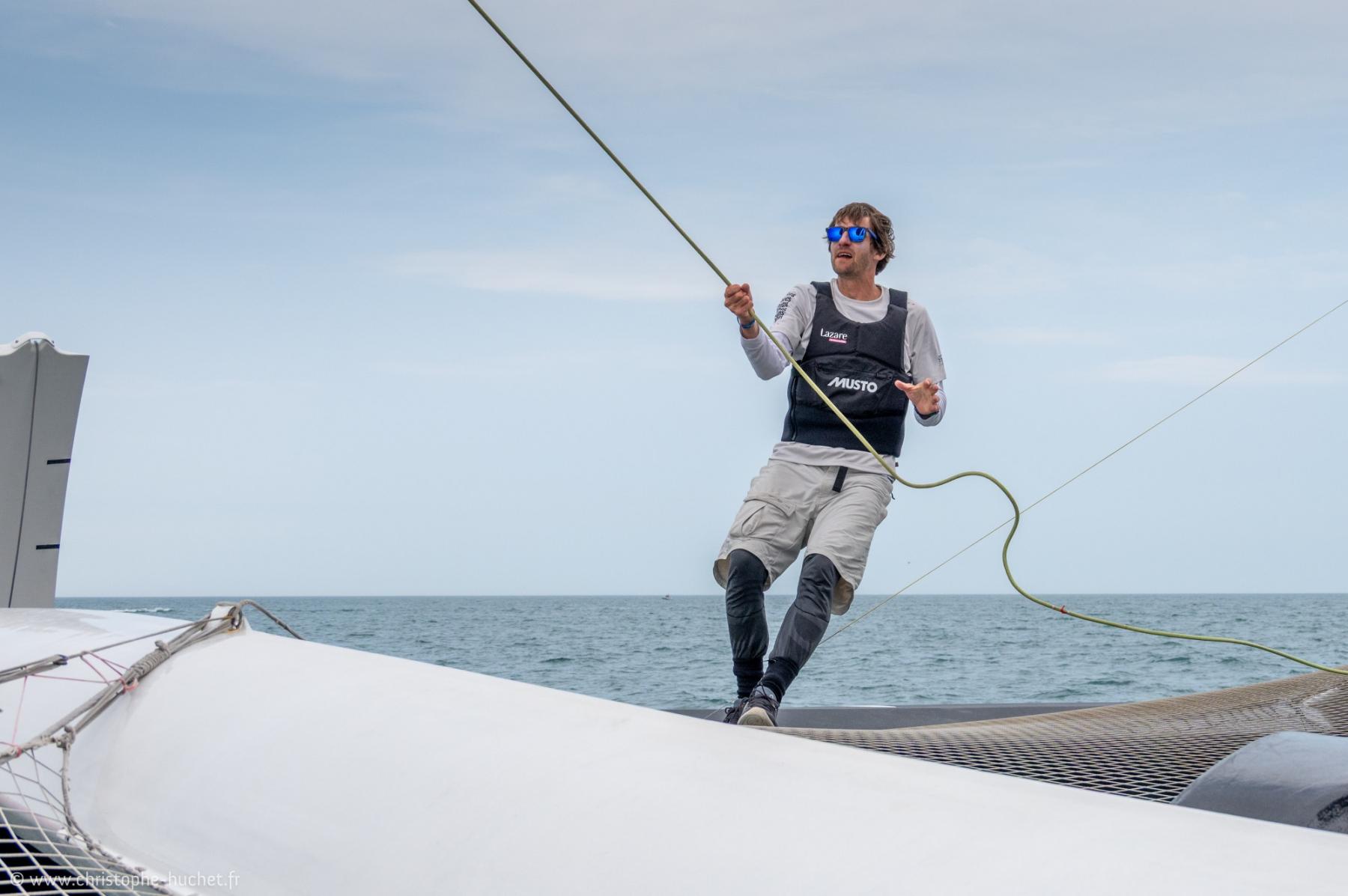 Marin sur un trimaran Ocean Fifty manipulant un cordage en mer sous un ciel clair lors d’une course à Saint-Malo.