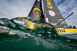 Voilier Imoca Café Joyeux en mer à Lorient, vue au ras de l’eau avec bulles et vagues sous un ciel clair.