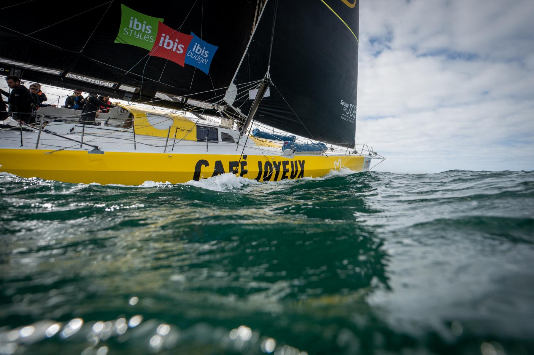 Coque jaune du voilier IMOCA Café Joyeux fendant les vagues à fleur d’eau lors d’une course au large.