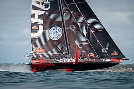 Voilier IMOCA Charal noir et rouge volant sur ses foils à pleine vitesse au large de Lorient sous un ciel nuageux.