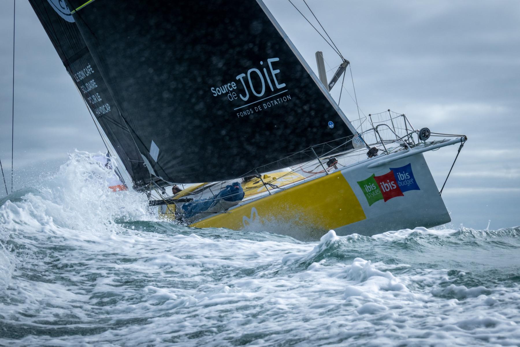 Voilier IMOCA jaune et blanc percutant une vague, voile noire Source de Joie, ciel nuageux et mer agitée.