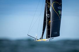 Vue au raz de l’eau du voilier IMOCA Café Joyeux, aux voiles noires et logo coeur jaune naviguant sur l’océan à Lorient sous un ciel bleu.