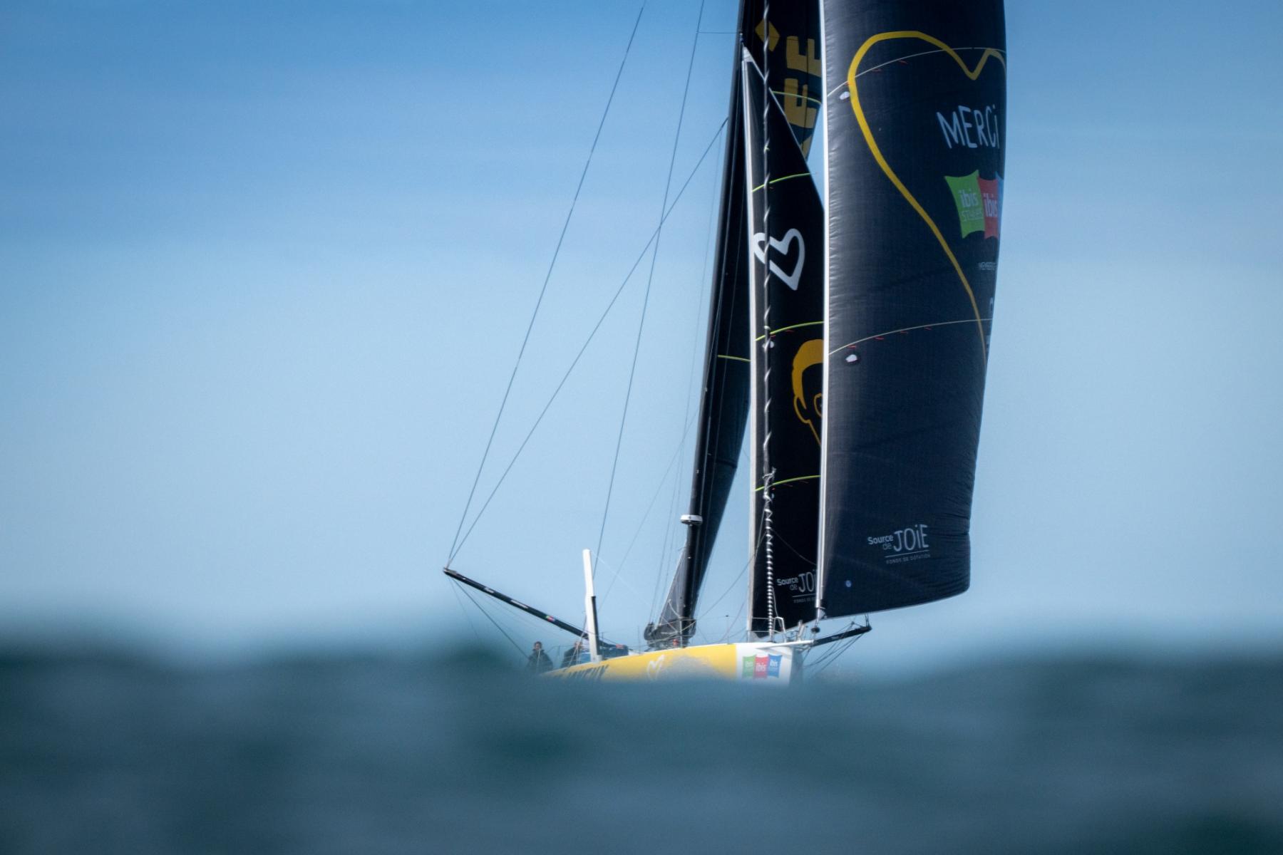 Vue au raz de l’eau du voilier IMOCA Café Joyeux, aux voiles noires et logo coeur jaune naviguant sur l’océan à Lorient sous un ciel bleu.