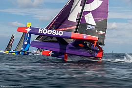 Trimaran Ocean Fifty Koesio violet et orange fendant les vagues à Lorient lors d’une régate nautique.