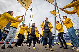 Équipiers en vestes jaunes célébrant avec drapeaux devant un voilier, contre-plongée, ambiance festive au port du Havre.