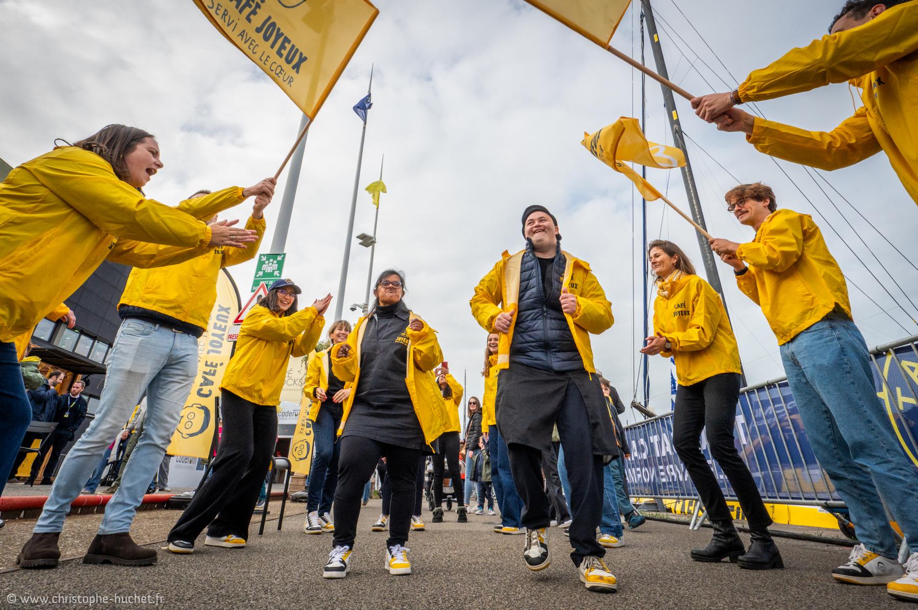Équipiers en vestes jaunes célébrant avec drapeaux devant un voilier, contre-plongée, ambiance festive au port du Havre.
