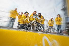 Équipe en cirés jaunes sabrant le champagne sur le pont d’un voilier Imoca jaune au Havre sous une lumière vive.