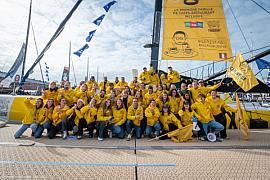 Groupe nombreux en vestes jaunes devant le mât d’un voilier Imoca sur un ponton au Havre sous un ciel clair.