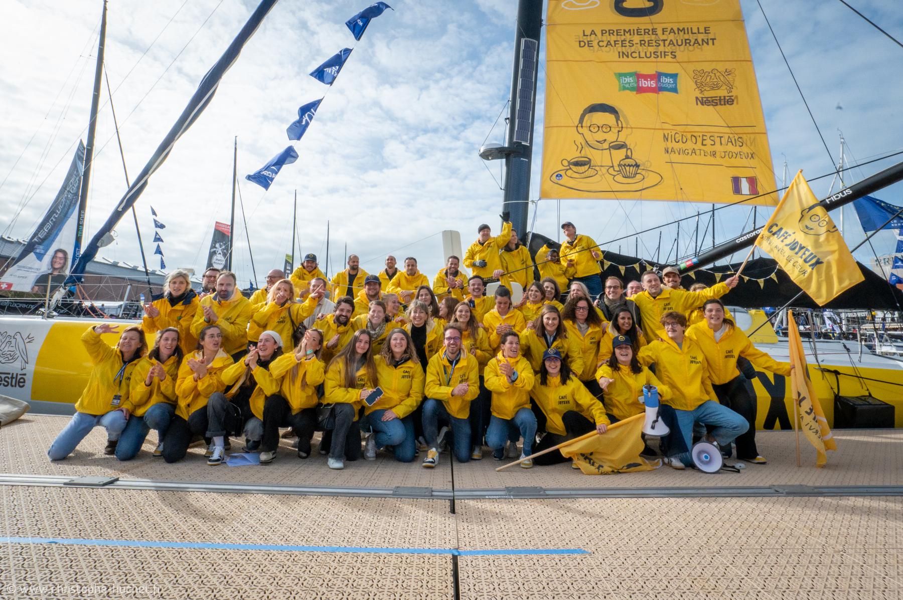 Groupe nombreux en vestes jaunes devant le mât d’un voilier Imoca sur un ponton au Havre sous un ciel clair.