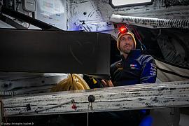 Skipper Simon Koster en cabine IMOCA avec lampe frontale rouge, intérieur technique de course et ambiance nocturne.