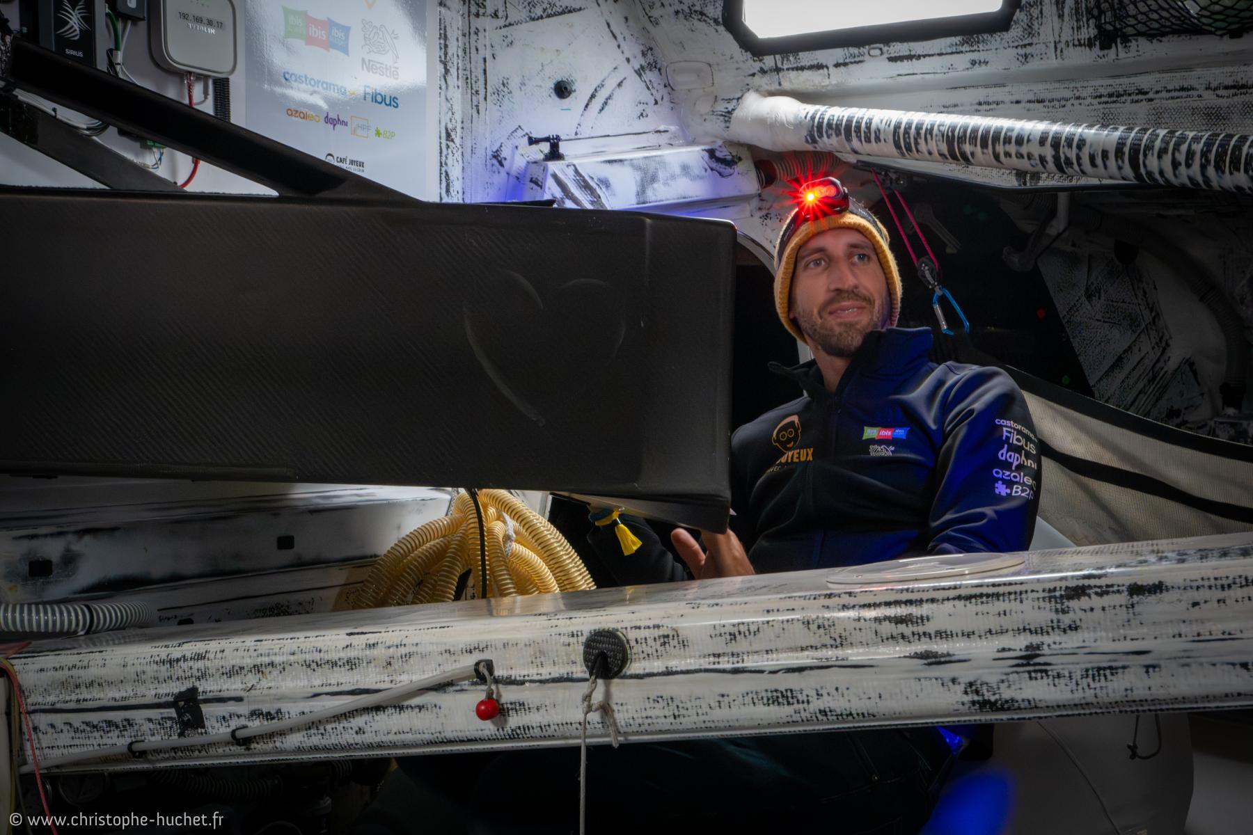 Skipper Simon Koster en cabine IMOCA avec lampe frontale rouge, intérieur technique de course et ambiance nocturne.