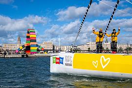 Skippeurs en vestes jaunes saluant sur un voilier Café Joyeux devant la Catène de containers colorés au Havre