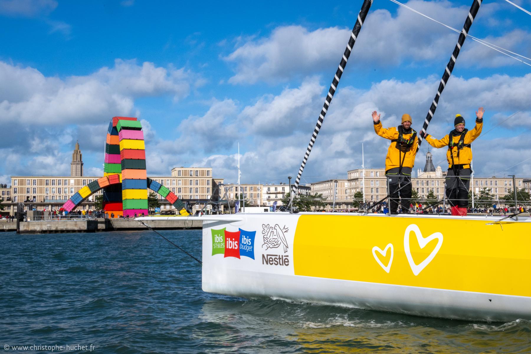 Skippeurs en vestes jaunes saluant sur un voilier Café Joyeux devant la Catène de containers colorés au Havre