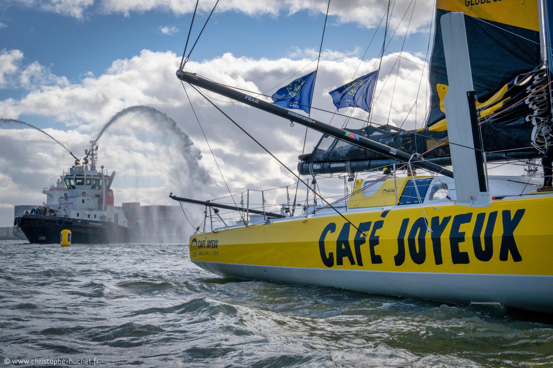 Coque jaune du voilier Café Joyeux au premier plan et remorqueur avec jets d’eau en arrière-plan au Havre