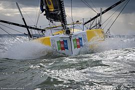 Imoca jaune et blanc Café Joyeux vu de face, fendant les vagues au Havre sous un ciel gris, écume et vitesse.