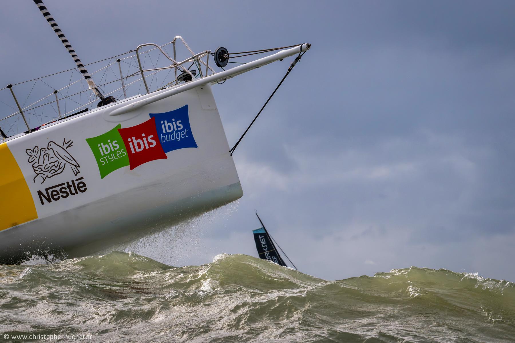 Étrave blanche de voilier avec logos Nestlé et Ibis fendant les vagues sous un ciel gris menaçant au Havre.