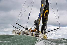 Skipper Nico d’Estais barrant son voilier IMOCA noir et jaune Café Joyeux sur une mer agitée sous un ciel nuageux.
