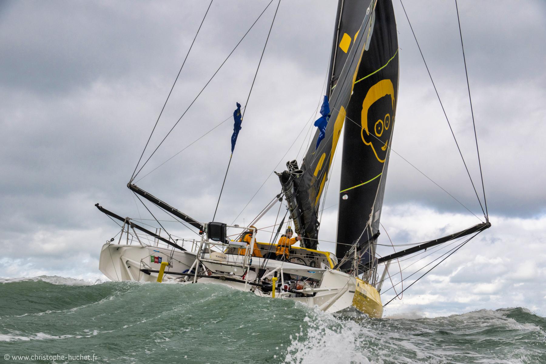 Skipper Nico d’Estais barrant son voilier IMOCA noir et jaune Café Joyeux sur une mer agitée sous un ciel nuageux.