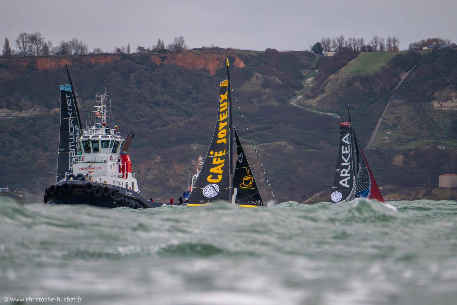 Remorqueur et voiliers Imoca Café Joyeux et Arkea en mer agitée devant les falaises du Havre sous un ciel gris.
