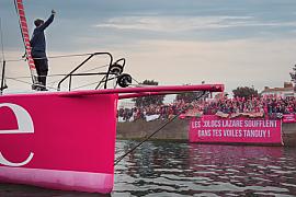 Skipper saluant la foule sur l’étrave rose du voilier Lazare, banderole de supporters en arrière-plan.